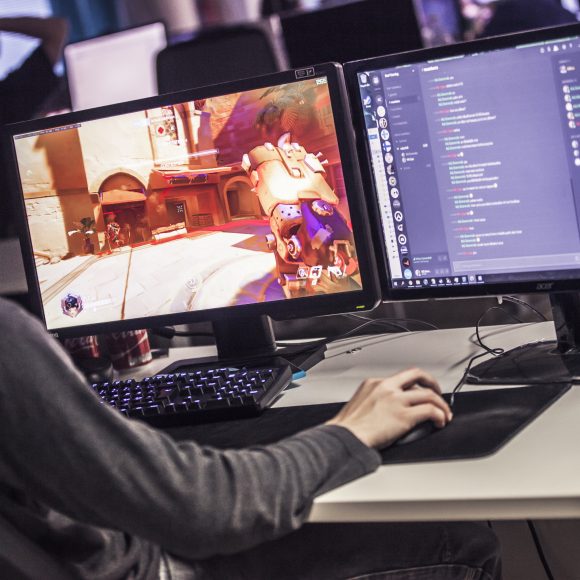 Henkilö pelaa tietokonepeliä kahden tietokonenäytön edessä.