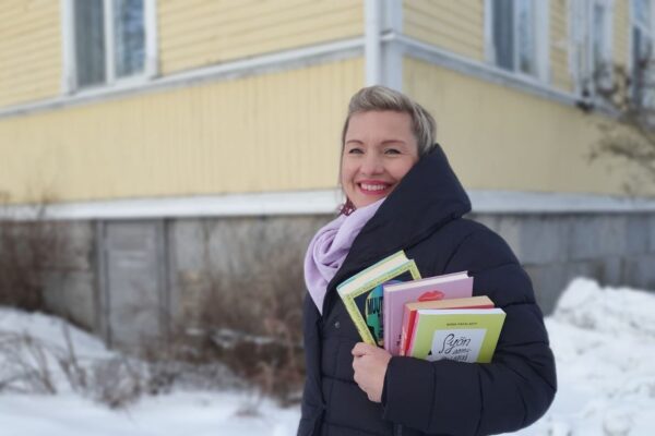 Helena Murtonen-Leppä seisoo hymyillen, kirjoja kädessään keltaisen vanhan puutalon pihalla.