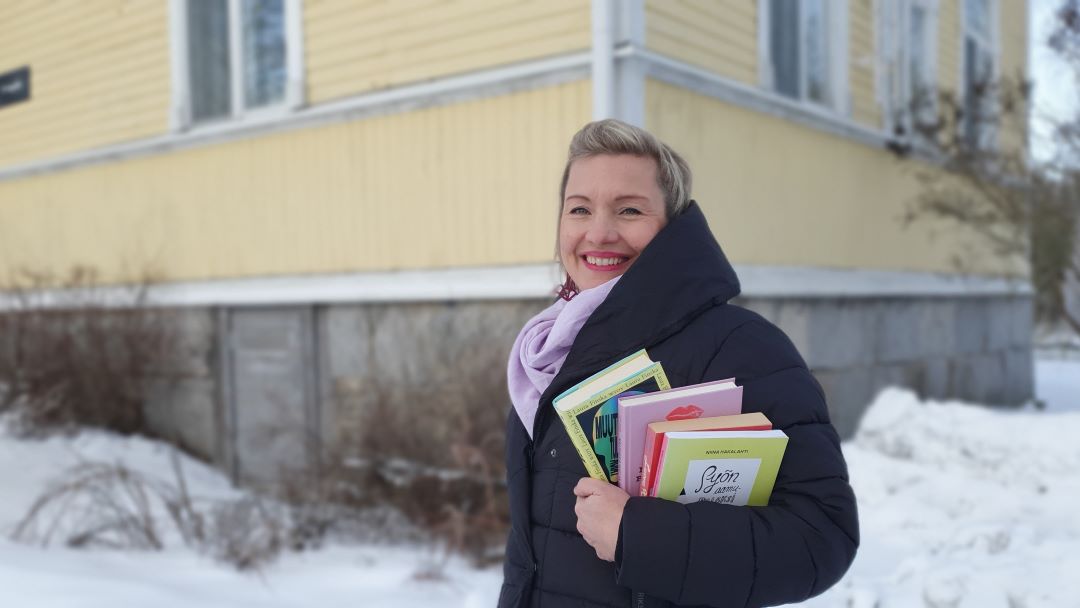 Helena Murtonen-Leppä seisoo hymyillen, kirjoja kädessään keltaisen vanhan puutalon pihalla.
