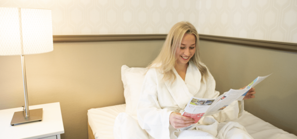 Nainen lukee hotellihuoneen sängyllä esitettä kylpytakki päällä.