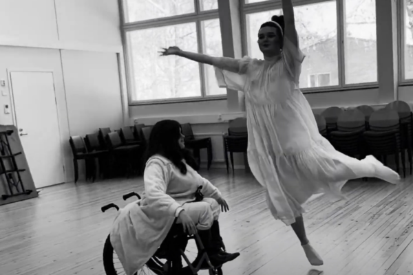 Henkilö istuu pyörätuolissa valkeissa vaatteissa toisen henkilön tanssiessa valkoinen mekko päällään hänen ympärillään.